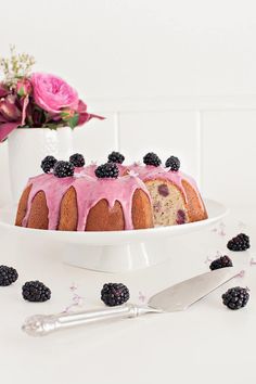 a bundt cake with pink icing, blackberries and flowers on the side