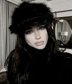 a woman with long hair wearing a fur hat and black coat in front of a mirror