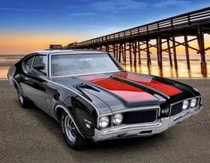 an old muscle car parked in front of the ocean with a pier in the background