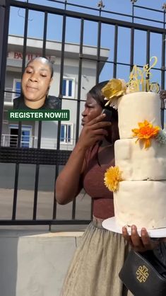 a woman holding a large white cake with yellow flowers on it in front of a gate