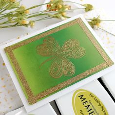 some type of green and gold card on a white surface with flowers in the background