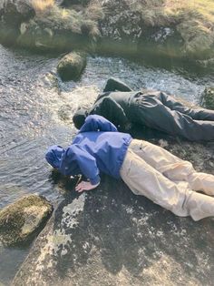 two people laying on rocks next to a body of water with grass and trees in the background