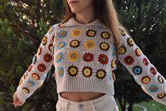 a woman standing in front of trees wearing a crocheted sweater with flowers on it