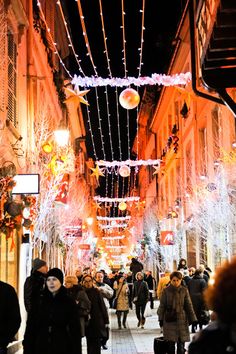 many people are walking down the street with lights on all over them and decorations hanging from the buildings