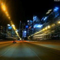 a car driving down a city street at night with bright lights on the buildings in the background