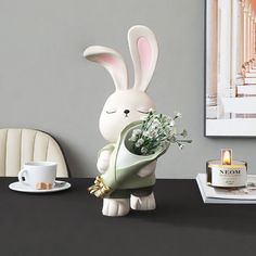 a white bunny holding a bouquet of flowers on a table next to a cup and saucer