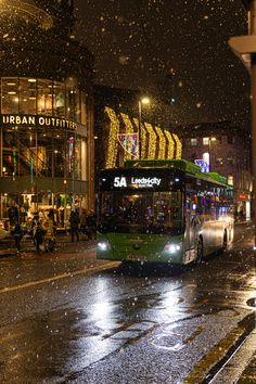 a green bus driving down a street next to tall buildings covered in snow at night