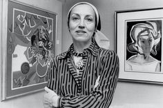 an older woman standing in front of some art on the wall with her arms crossed