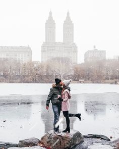 two people standing on top of a rock next to a body of water with buildings in the background