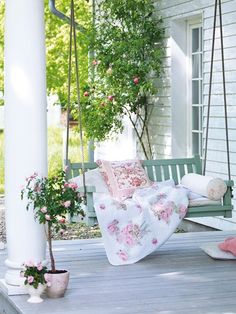 a white porch swing with pink flowers and pillows on it, next to a potted plant