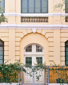 a yellow building with green shutters and a white door on the outside, surrounded by greenery