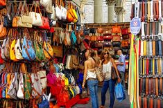 two women walk through an outdoor market selling handbags and purses