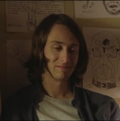 a man with long hair standing in front of a wall full of drawings and pictures