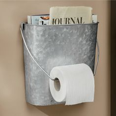 a metal bucket holding toilet paper and a roll of tissue hanging on the wall next to a magazine rack