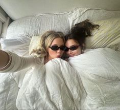 two young women laying in bed with white sheets and blankets on top of each other