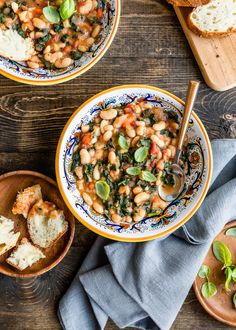 two bowls filled with beans and spinach on top of a wooden table next to bread