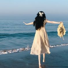 a woman in a dress and hat walking on the beach with her arms spread out