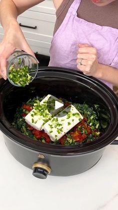 a woman in an apron is pouring something into a crock pot filled with vegetables