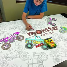 a young boy sitting at a table with monster trucks on it and coloring the paper