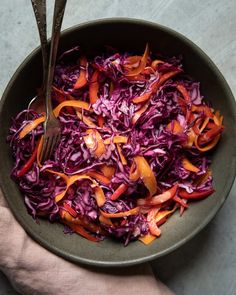 a bowl filled with red cabbage and carrots on top of a table next to a person's hand