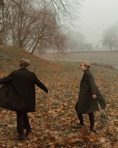 two people walking through leaves in the middle of an open field on a foggy day