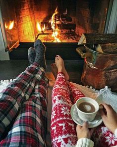 two people sitting in front of a fireplace with their feet up on a plate and holding a cup of coffee