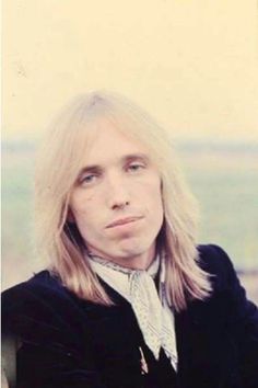 a man with long blonde hair wearing a black jacket and white shirt looking off into the distance