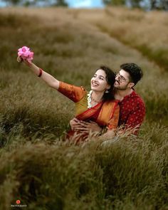 a man and woman are sitting in the tall grass holding a pink flower up to their face