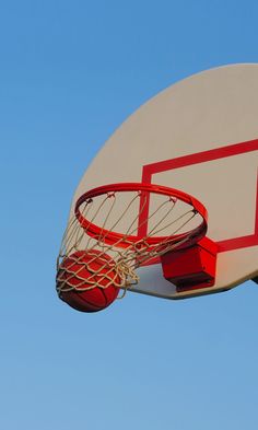 a basketball going through the rim of a white and red hoop on a clear day