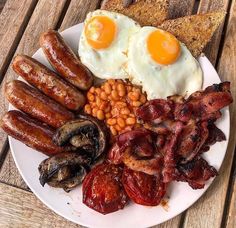 an english breakfast with bacon, eggs, beans and toast on a white plate sitting on a wooden table