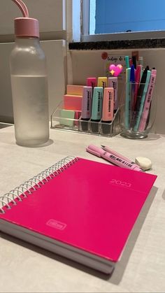 a pink notebook and pen sitting on a table next to a bottle with pens in it