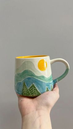 a hand holding a coffee mug with mountains and trees painted on the inside, in front of a gray background