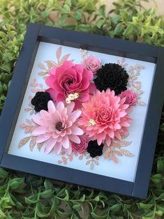 pink and black paper flowers are in a shadow box on green grass with gold leaves