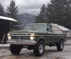 an old green truck parked in front of a barn on a snowy day with mountains in the background