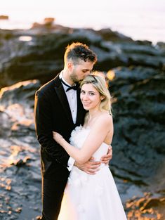 a man and woman standing next to each other in front of the ocean at sunset