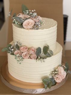 a three layer white cake with pink flowers and greenery on the top, sitting on a wooden table