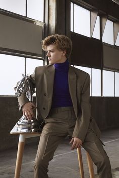 a young man sitting on top of a wooden chair wearing a suit and turtle neck sweater