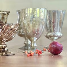 an assortment of glass goblets sitting on a wooden table with fruit in the foreground