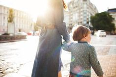 a woman holding the hand of a little boy walking down a street with buildings in the background