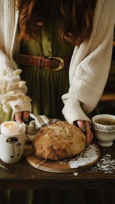 a woman holding a loaf of bread on top of a wooden table next to cups and saucers