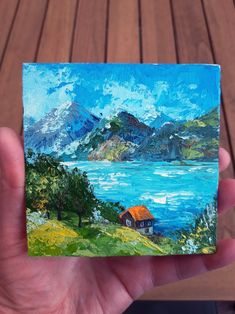 a hand holding up a small painting of a house by the water with mountains in the background