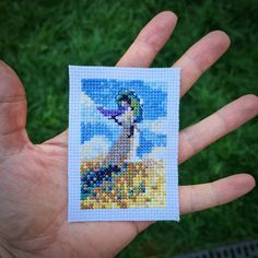 a hand holding a small cross - stitch picture of a woman in the sky and clouds