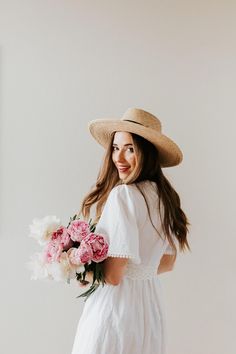 a woman in a white dress and straw hat holding a bouquet of pink peonies