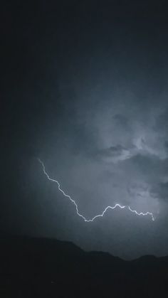 a lightning bolt is seen in the dark sky above some hills and trees on a cloudy day