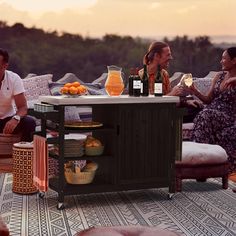 three people sitting around a table with drinks and food on it while the sun is setting