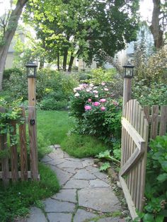 an open gate leading to a lush green yard with flowers and trees in the background