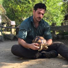 a man sitting on the ground petting a tortoise in front of him