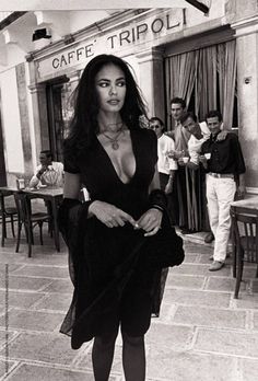 a black and white photo of a woman standing in front of a cafe with people behind her