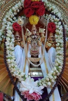 the statue is surrounded by flowers and garlands on it's head, in front of a gold plated backdrop