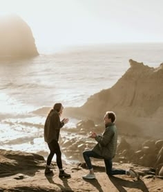 two people sitting on rocks near the ocean and one is holding something in his hand